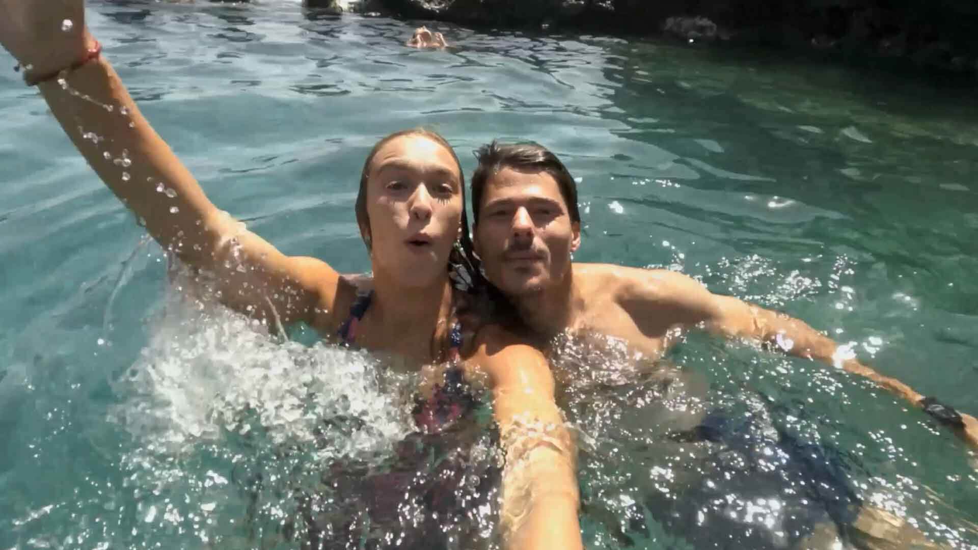 Dos personas bañándose en agua cristalina, con una de ellas levantando el brazo y rocas al fondo.