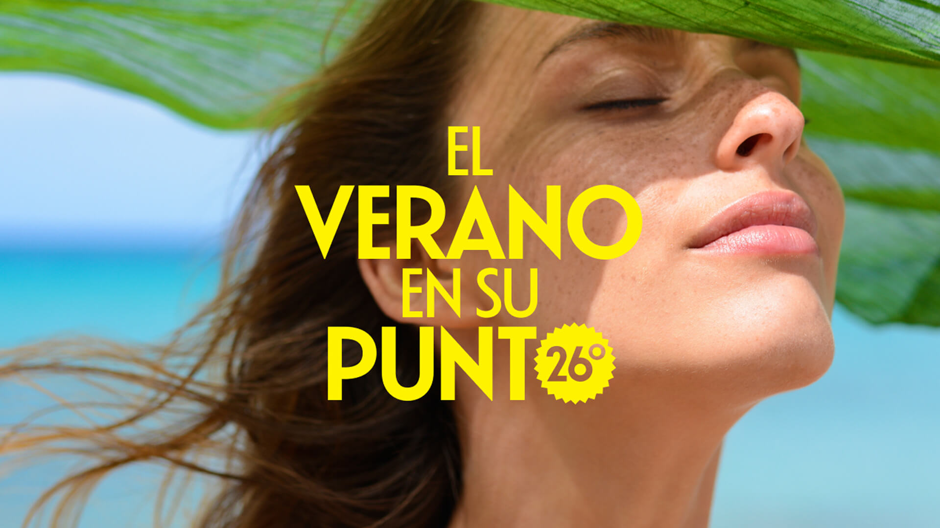 Persona con los ojos cerrados y el rostro al sol, con una tela verde moviéndose sobre la cabeza y el cabello al viento, con el mar al fondo.