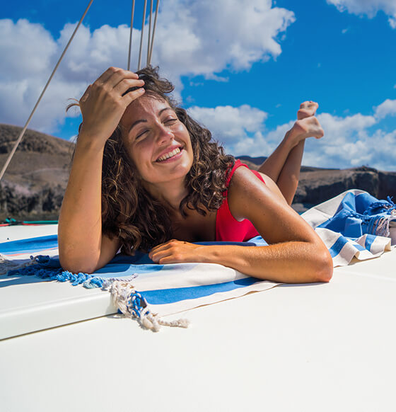 Persona descansando sobre la cubierta de un velero en el mar, con acantilados al fondo en un día soleado.
