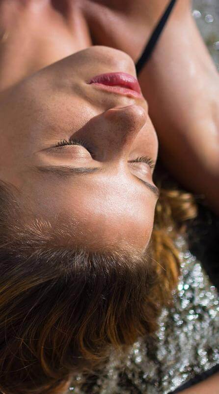 Persona tumbada con los ojos cerrados tomando el sol sobre una superficie de piedra húmeda.