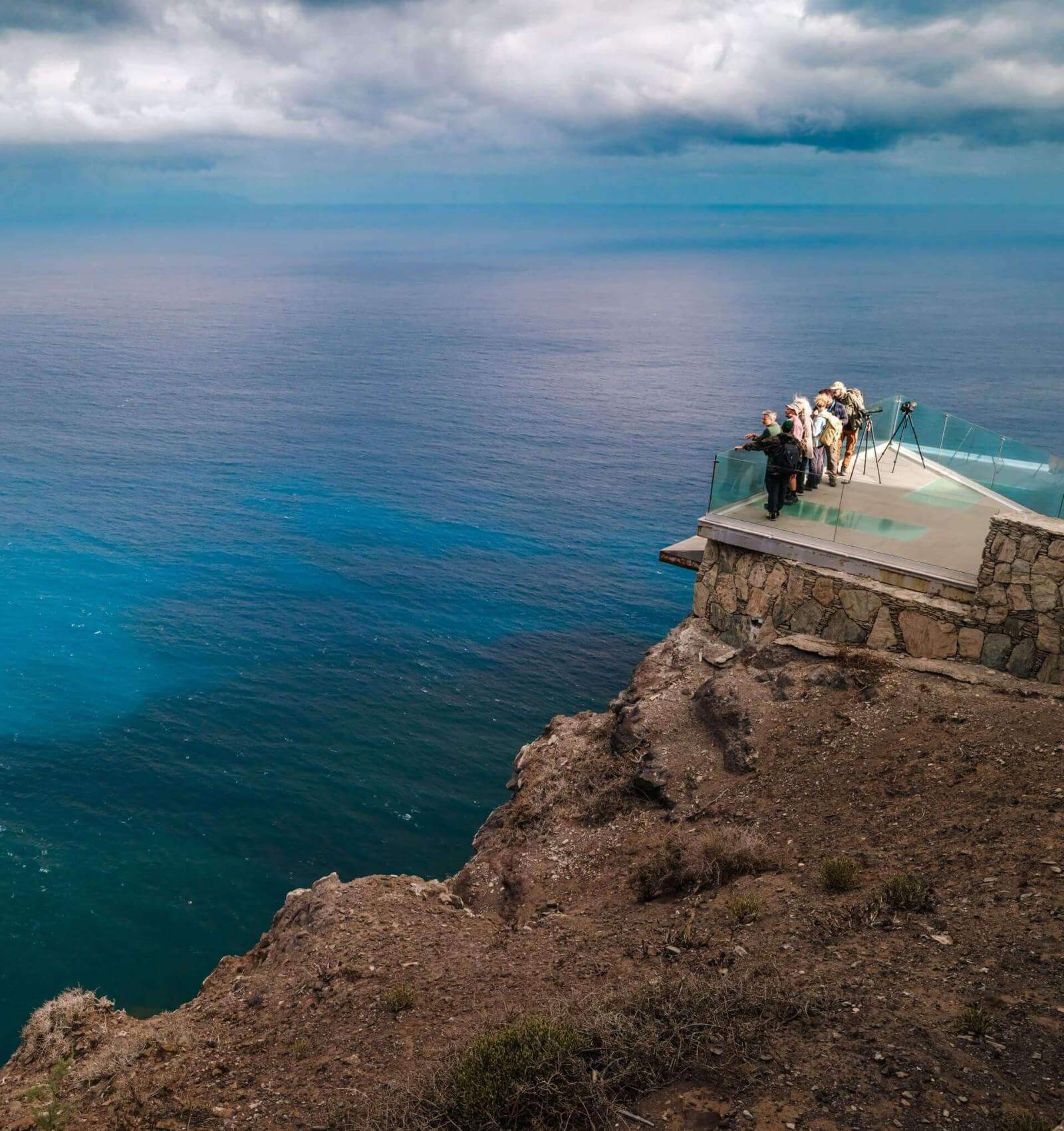 Personas observando el océano Atlántico desde mirador acristalado en acantilado bajo cielo nublado.
