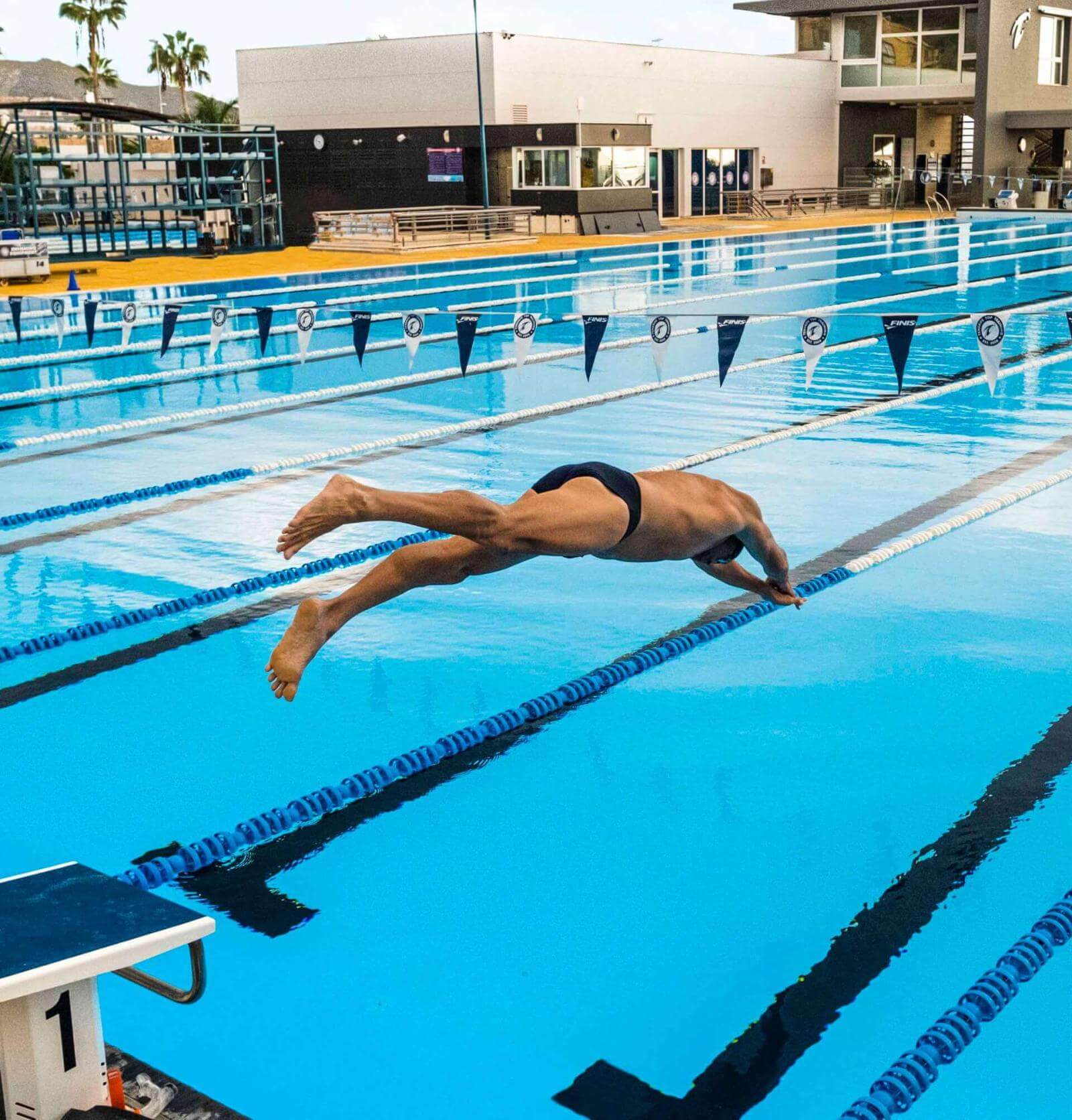 Persona realizando salto de entrada en piscina olímpica al aire libre con carriles y edificio deportivo al fondo.