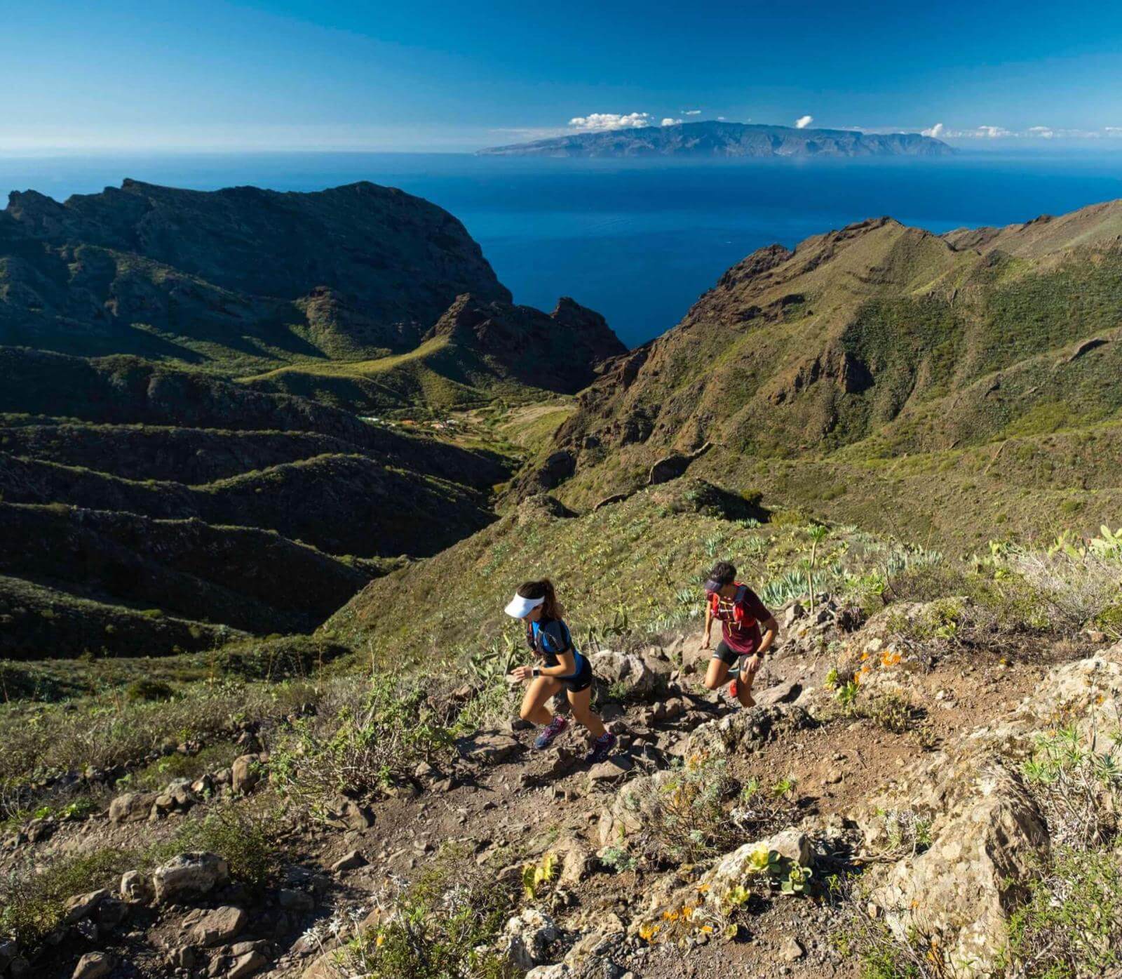 Dos personas practicando trail running en sendero montañoso con vistas al océano y otra isla en el horizonte.