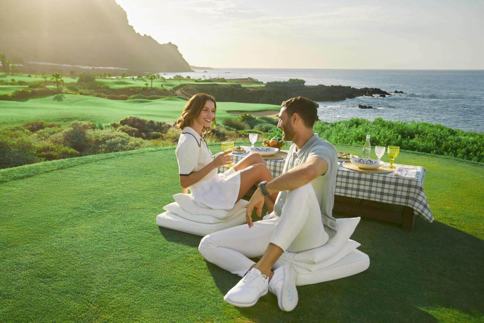 Pareja disfrutando de un picnic con mesa servida en campo de golf frente al océano y montañas en Islas Canarias.