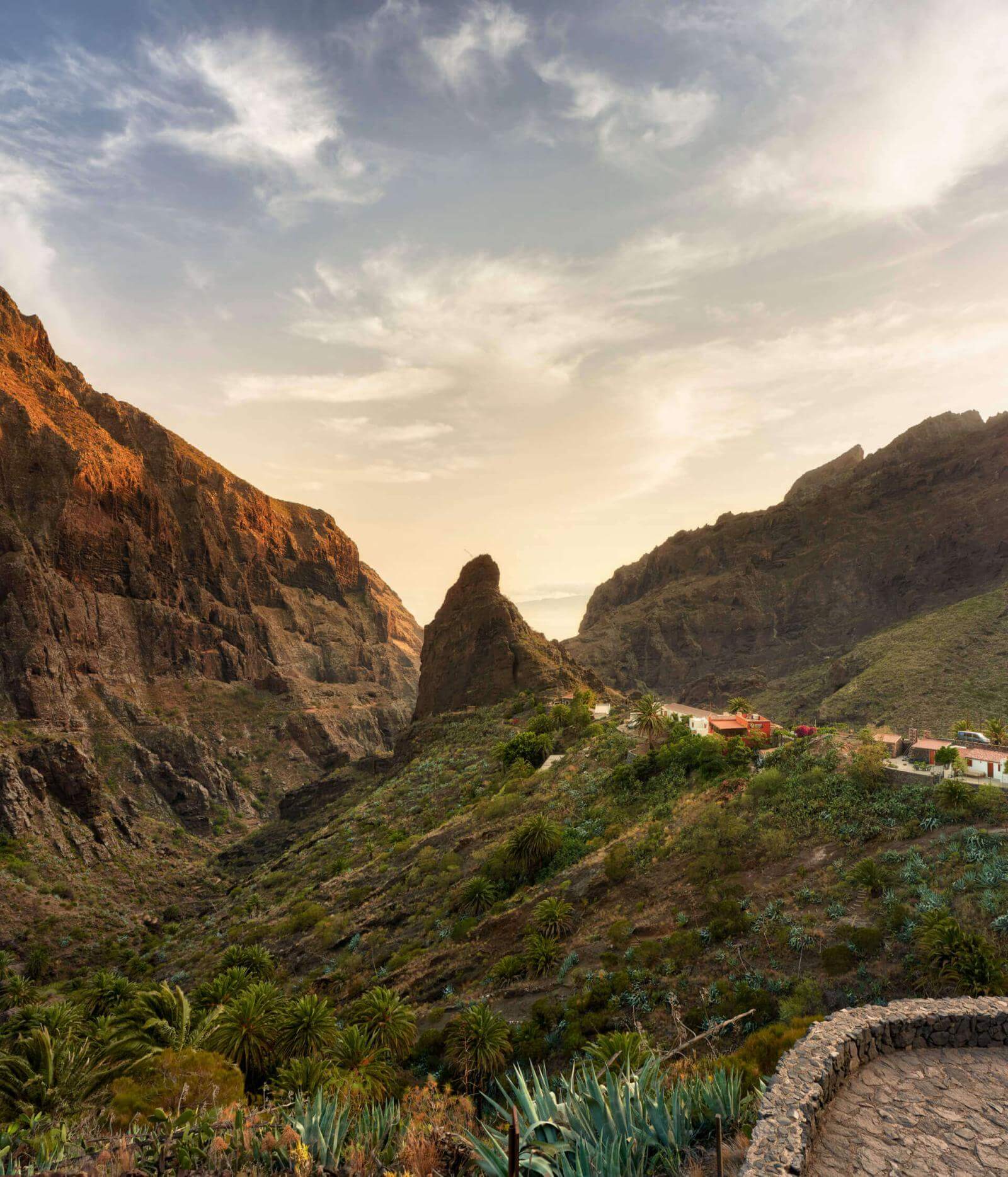 Acantilados rocosos en un valle con vegetación y casas en la ladera, iluminado por la suave luz del atardecer.