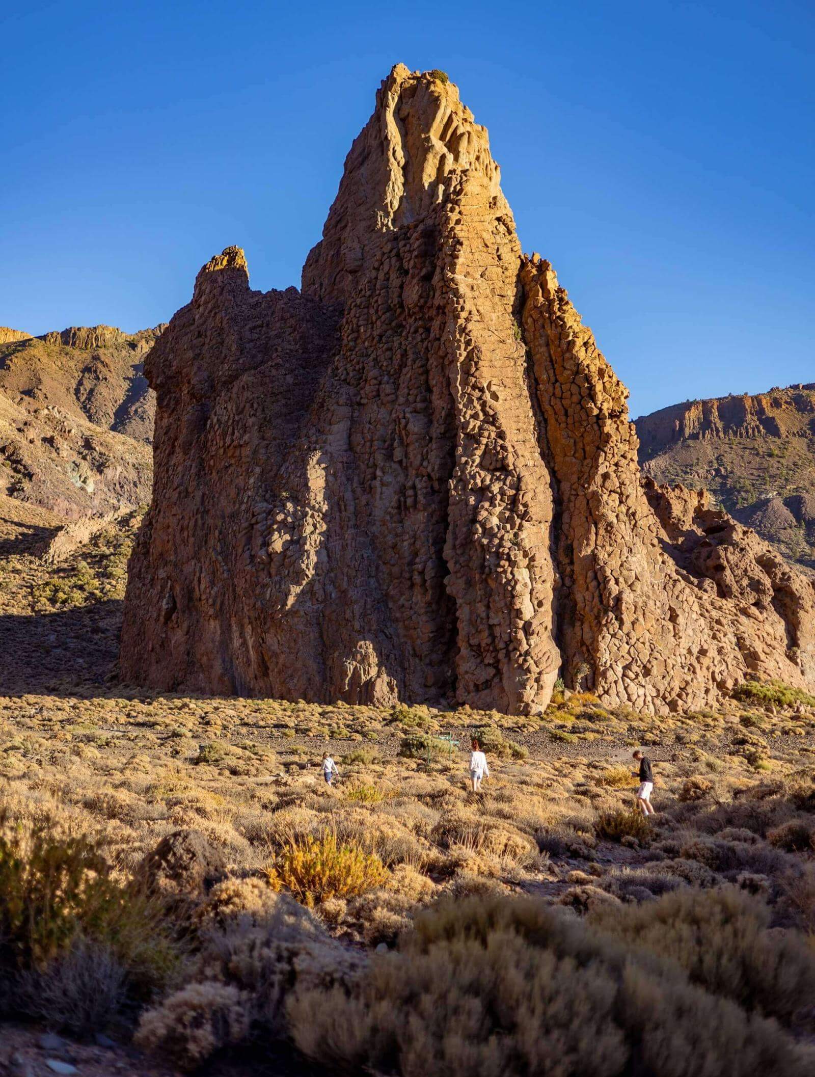 Personas caminando entre matorrales bajos frente a una gran formación rocosa vertical en un paisaje volcánico.