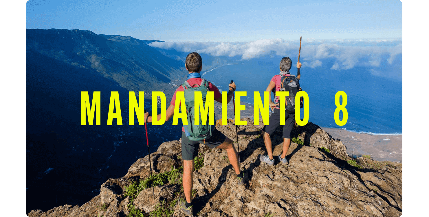 Dos personas con mochilas en lo alto de un acantilado, observando un valle verde y la costa con nubes bajas.
