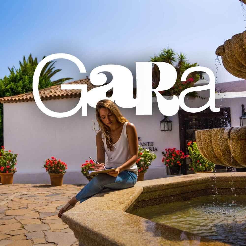 Persona escribiendo en un cuaderno junto a una fuente de piedra en un patio con flores y edificio de arquitectura tradicional. 