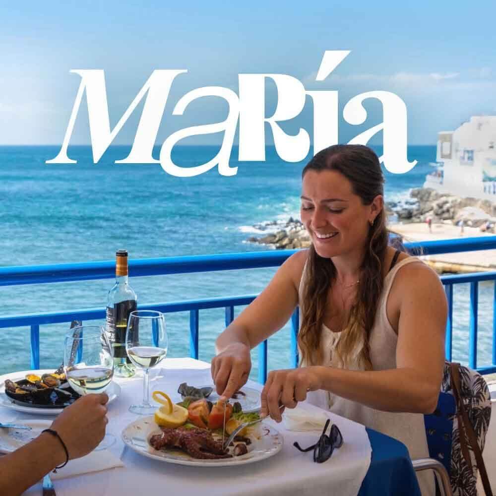 Persona disfrutando de una comida en terraza con vistas al mar, con platos de marisco y copas de vino. 