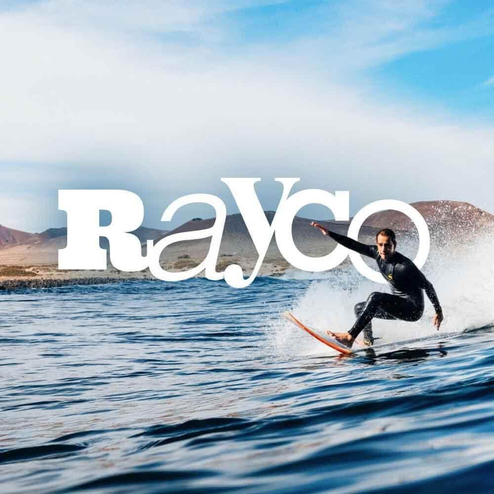 Persona practicando surf sobre una ola en el mar, con salpicaduras de agua y paisaje volcánico al fondo. 