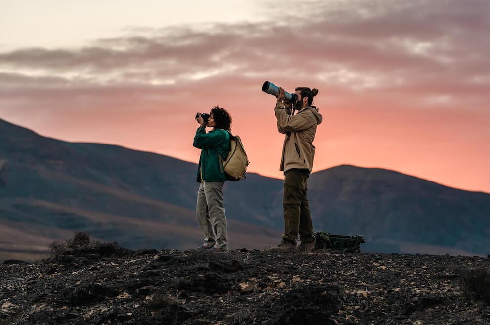Dos personas observando aves con prismáticos y teleobjetivo en paisaje volcánico al amanecer.