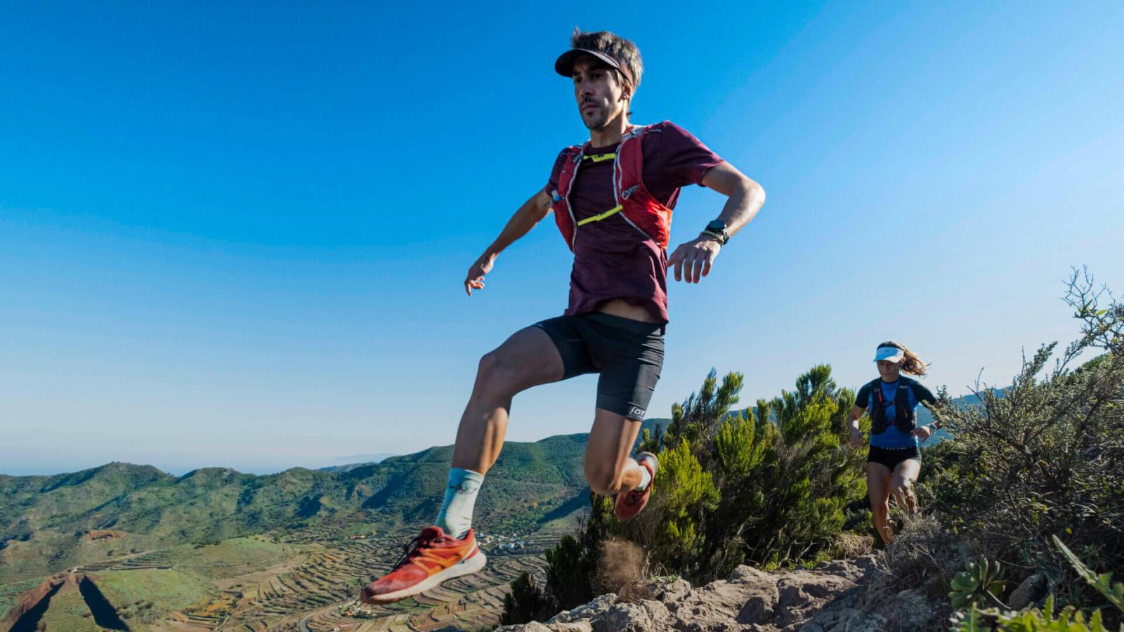 Personas corriendo por sendero rocoso en entorno natural montañoso con vegetación y cielo despejado.