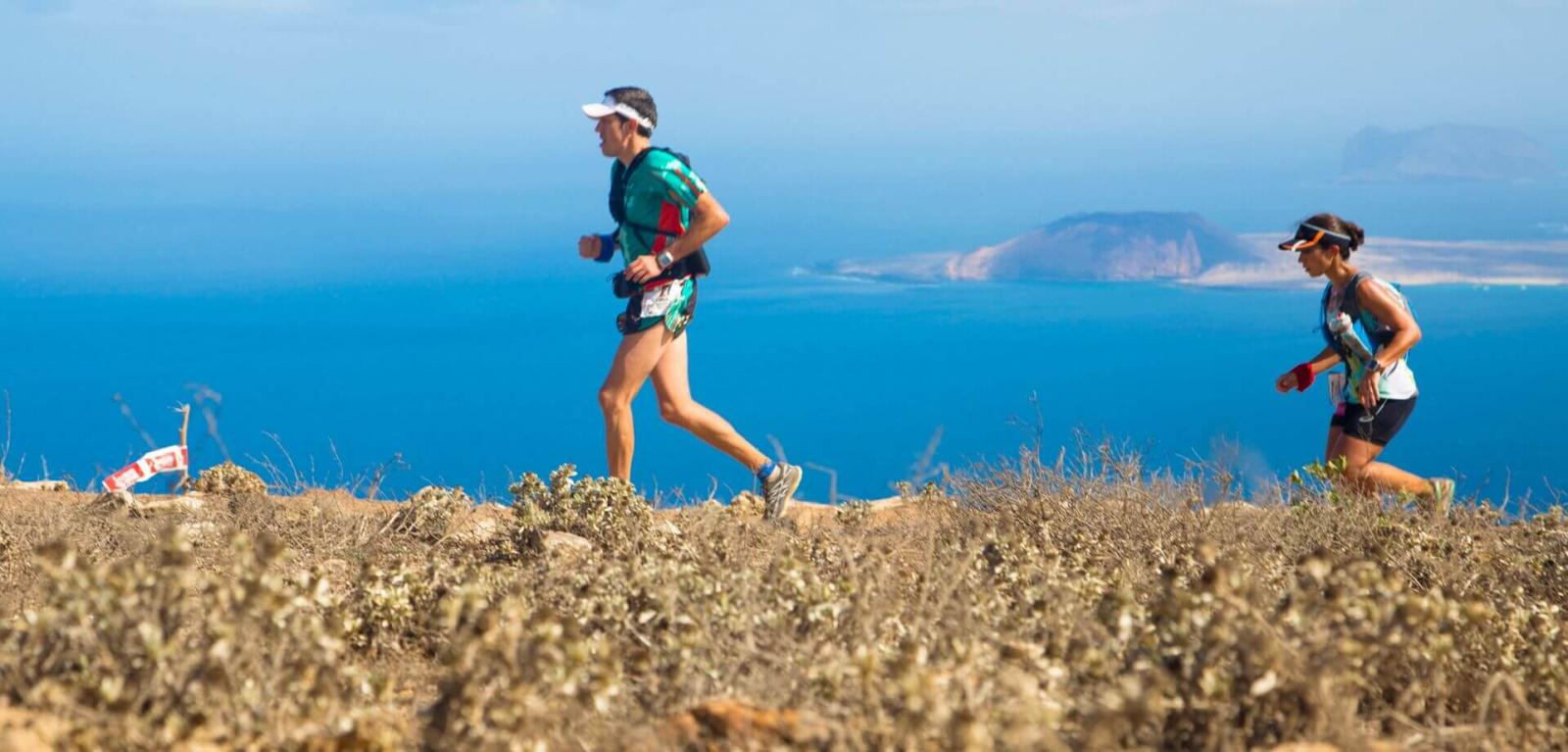 Pareja de atletas entrenando en sendero montañoso con paisaje volcánico y el mar de fondo.