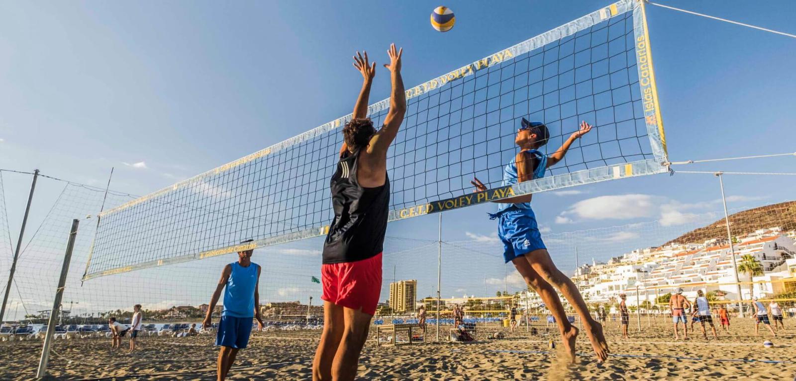 Personas practicando vóley playa y saltando para disputarse un balón.