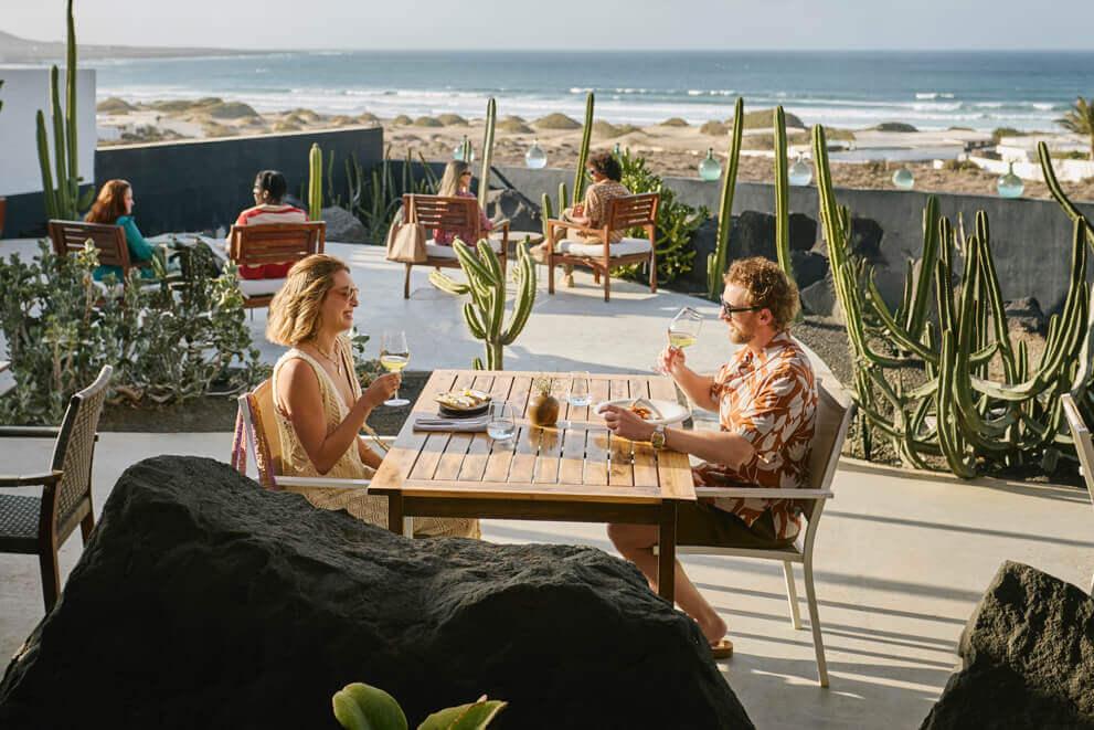 Dos personas sentadas en terraza al aire libre disfrutando de comida y bebida, con cactus y vistas al océano.
