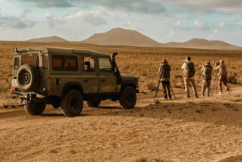 Grupo de personas con cámaras y trípodes observando aves en un paisaje árido junto a un todoterreno.