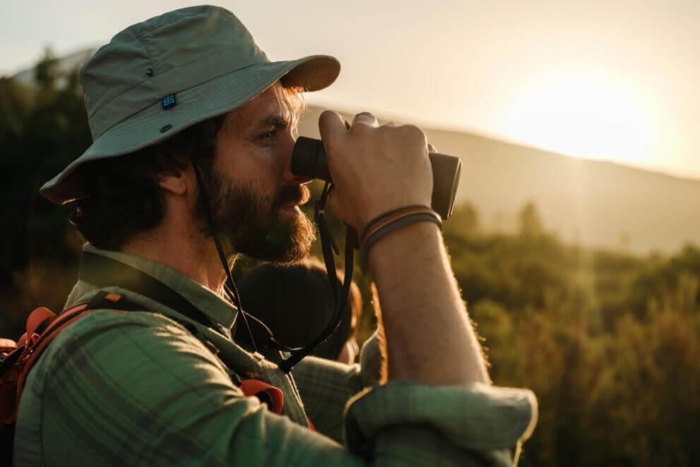 Primer plano de persona con sombrero de senderismo observando el paisaje con unos prismáticos al atardecer.