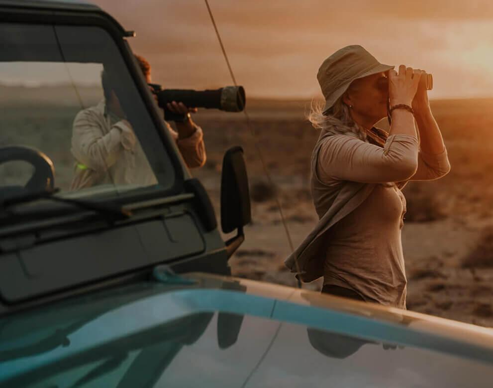Persona observando aves con prismáticos en un paisaje árido al atardecer junto a un vehículo todoterreno.