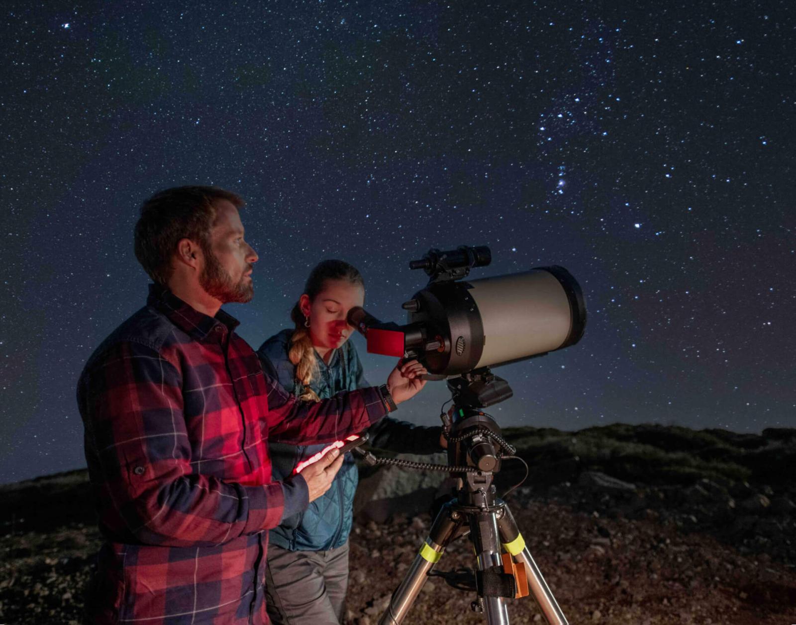 Dos personas usando un telescopio bajo un cielo nocturno estrellado en un paisaje rocoso.