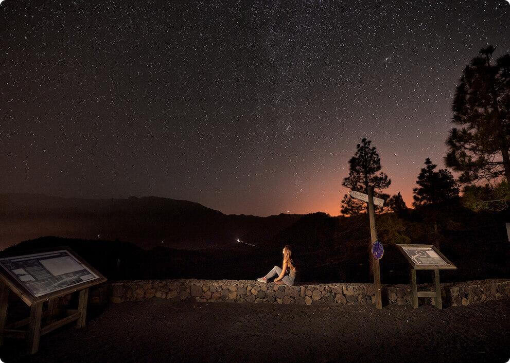 Noche estrellada desde el mirador Llano del Jable, en La Palma, con siluetas de montañas y una persona observando el paisaje.