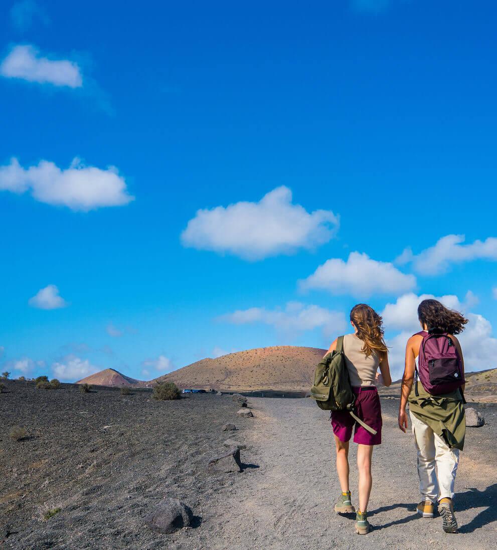 Dos personas caminando por un sendero volcánico en el Parque Nacional de Timanfaya bajo cielo azul.