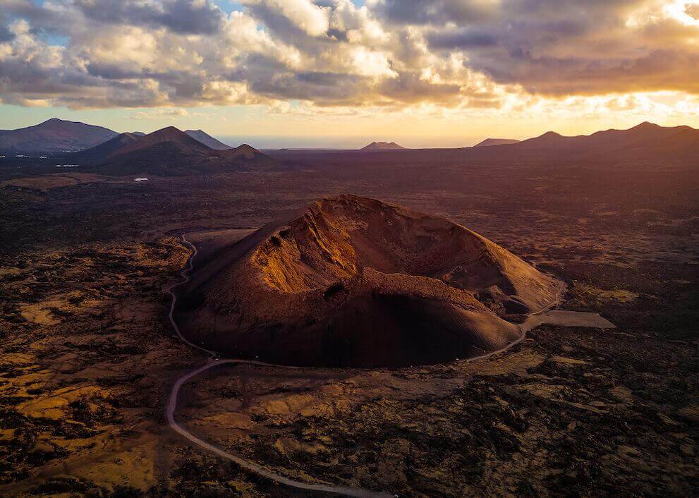 Vista aérea del Volcán del Cuervo en Lanzarote, con su cráter iluminado al atardecer y un paisaje volcánico alrededor.