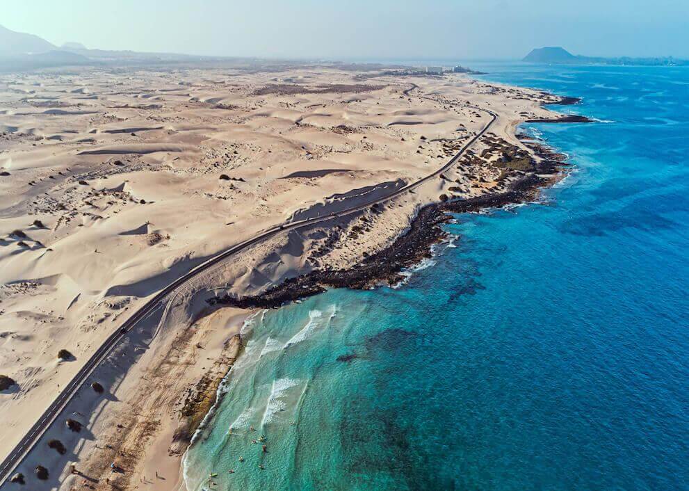 Vista aérea de las Dunas de Corralejo en Fuerteventura, con extensos arenales junto al mar turquesa y la costa volcánica.