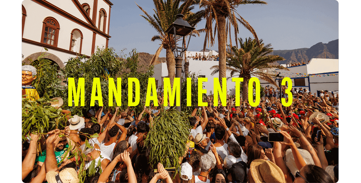 Multitud celebrando una festividad al aire libre entre palmeras y edificios tradicionales, levantando ramos verdes.