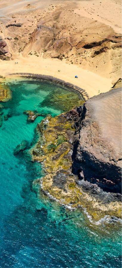 Vista aérea de la playa del Papagayo en Lanzarote, con arena dorada, acantilados volcánicos y mar turquesa.