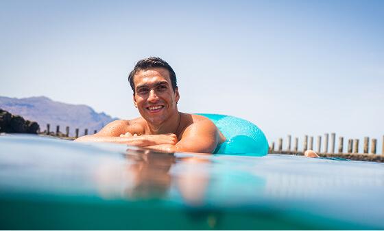 Persona apoyada sobre un flotador en una piscina natural, con superficie tranquila y postes de madera al fondo.
