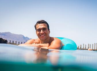 Persona apoyada sobre un flotador en una piscina natural, con superficie tranquila y postes de madera al fondo.