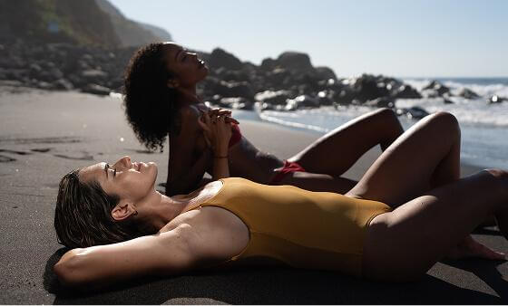 Dos personas tumbadas sobre arena oscura en una playa en un entorno rocoso.
